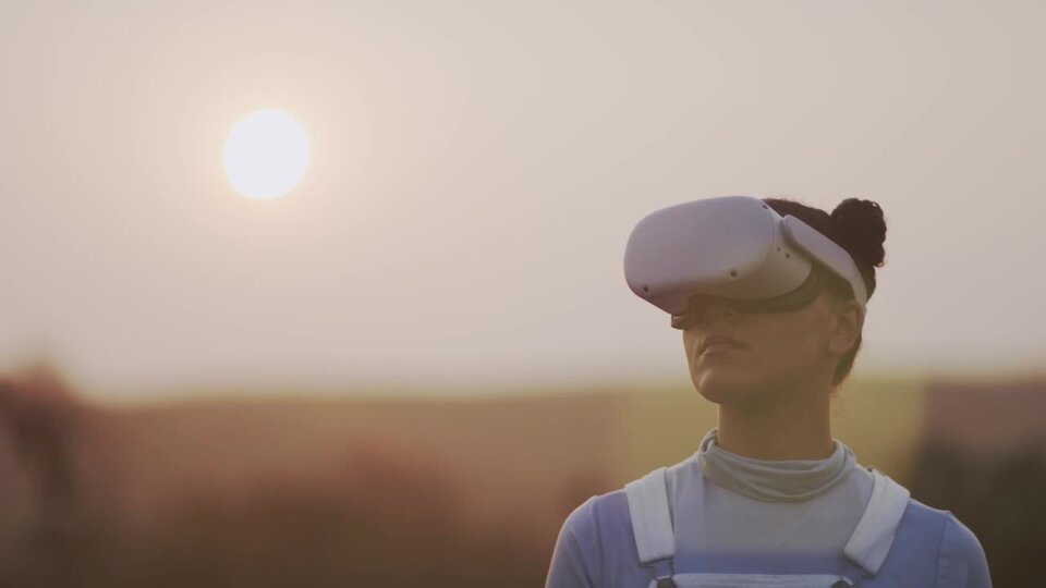 A young person wearing a white VR headset, light blue shirt, and white overalls gazes into the headset. The backgrounds shows the sun casting golden light over a prairie. The sunlight causes a lens flare over the person's face.