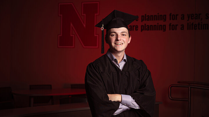 Business student Caleb Newburn stands, arms crossed, by a Hawks Hall wall decorated with the Nebraska N.