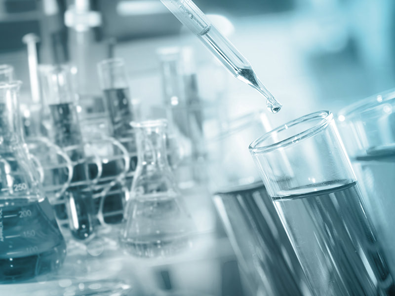 A pipette dropping liquid into a test-tube in a science laboratory, with more test tubes and beakers in the background. The whole scene has a light blue tint.