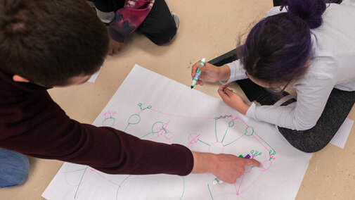 Aerial view of a youth and a young adult peering over a large piece of paper covered in network drawings. The youth holds a green marker in their hand, about to draw, as the young adult points to one of the networks on the paper.