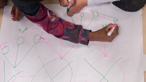 Hands of youth with brown skin draw pink and green network models on a large, white sheet of paper.