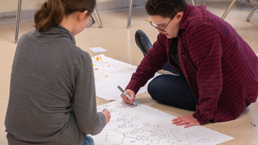 A youth and a young adult draw network models with markers on large sheets of white paper on the floor of a classroom.