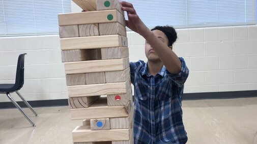 A young person with brown skin and dark, curly hair pulled back in an up-do, wearing a plaid flannel shirt and jeans, sits on a classroom floor. Their arms reach in front of their face to stack a wooden block on top of a giant tumbling tower.