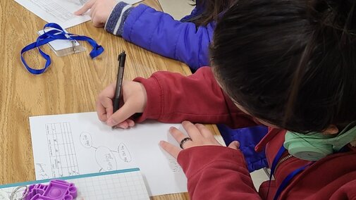 Two middle-school youth, one wearing purple and one wearing red, draw network maps on pieces of white paper.