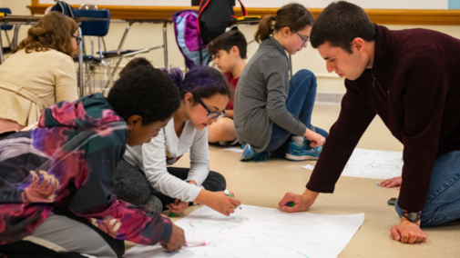 A young adult and youth sit on the floor of a classroom around a drawing of networks on a large piece of white paper. The youth hold markers and are talking about the drawing.
