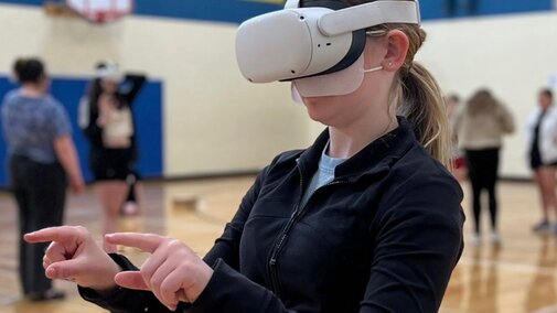 A young white person with blonde hair in a ponytail wearing a long sleeve black zipper-front shirt points forward with both fingers as they playtest a VR experience. They are wearing a white VR headset and standing in a middle school gym.