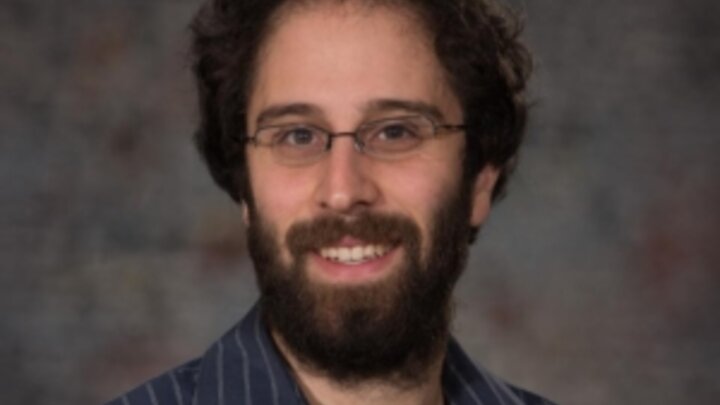 Headshot of a smiling person with dark hair, a beard, glasses, and a dark blue striped shirt