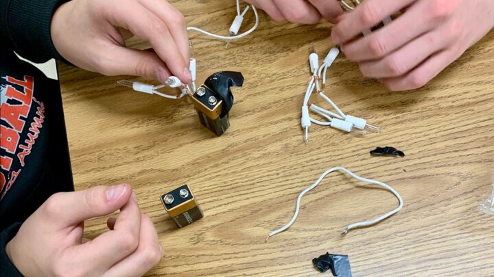 Two sets of light-skinned hands building simple circuits with incandescent lights, electrical tape, and 9-volt batteries