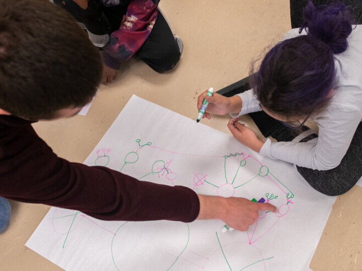 Aerial view of a youth and a young adult peering over a large piece of paper covered in network drawings. The youth holds a green marker in their hand, about to draw, as the young adult points to one of the networks on the paper.