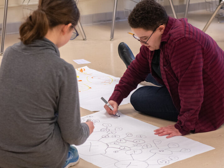 A youth and a young adult draw network models with markers on large sheets of white paper on the floor of a classroom.