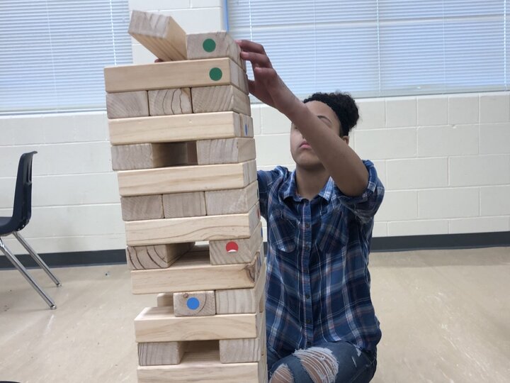 A young person with brown skin and dark, curly hair pulled back in an up-do, wearing a plaid flannel shirt and jeans, sits on a classroom floor. Their arms reach in front of their face to stack a wooden block on top of a giant tumbling tower.