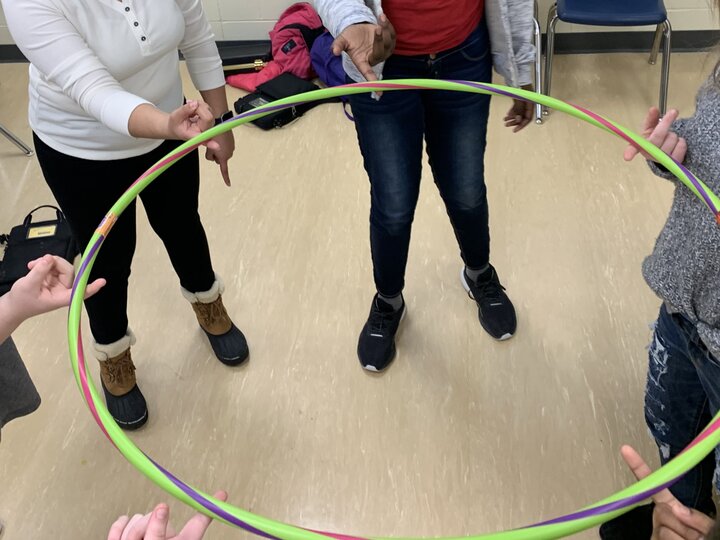 Hands of people with various skin tones balance a striped hula hoop on their index fingers in a classroom