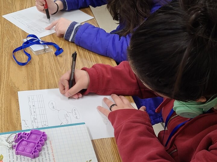 Two middle-school youth, one wearing purple and one wearing red, draw network maps on pieces of white paper.