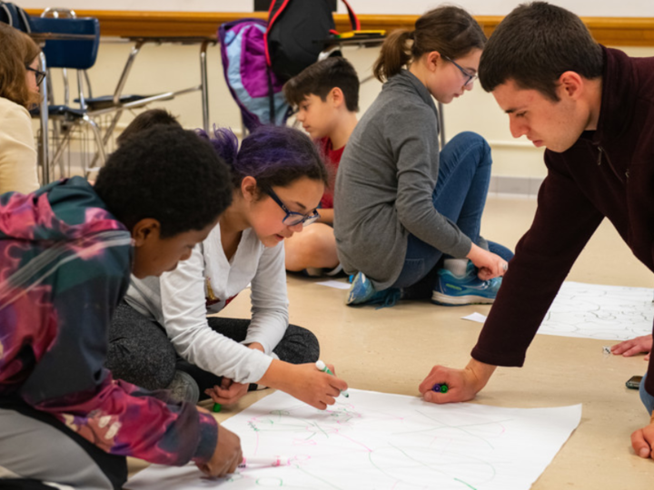 A young adult and youth sit on the floor of a classroom around a drawing of networks on a large piece of white paper. The youth hold markers and are talking about the drawing.