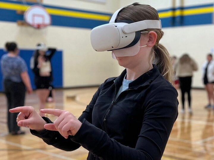 A young white person with blonde hair in a ponytail wearing a long sleeve black zipper-front shirt points forward with both fingers as they playtest a VR experience. They are wearing a white VR headset and standing in a middle school gym.