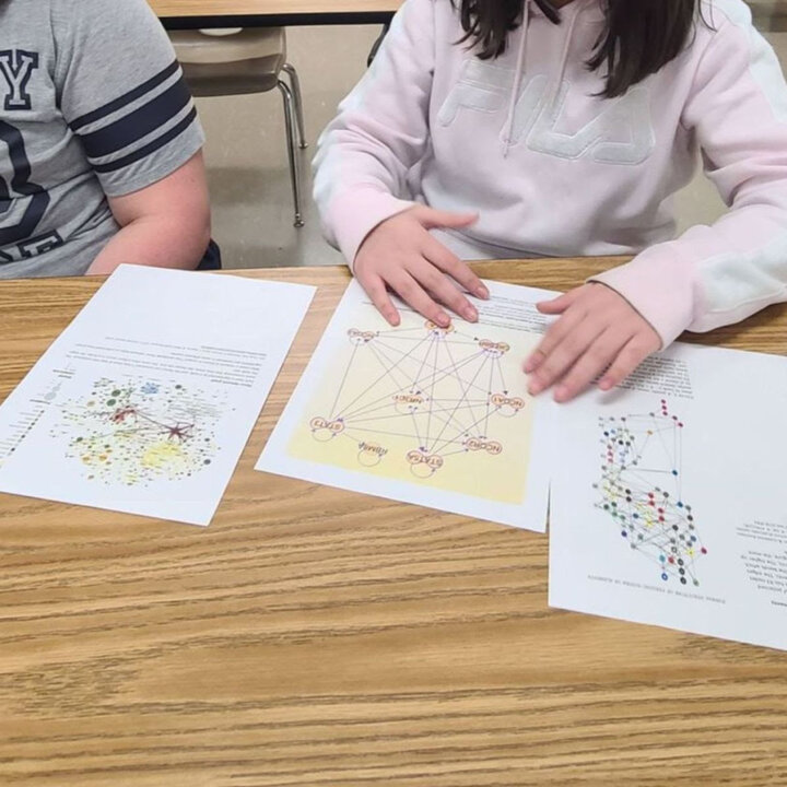 Network images spread out on a desk. Two youth with white skin sit at the desk and discuss the networks; you can see their torsos and arms.