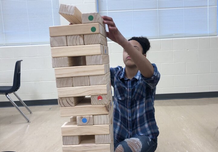 A young person with brown skin and dark, curly hair pulled back in an up-do, wearing a plaid flannel shirt and jeans, sits on a classroom floor. Their arms reach in front of their face to stack a wooden block on top of a giant tumbling tower.