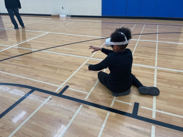 A young person with brown skin and black, curly hair sits on the floor of a middle school gym. They are wearing all-black clothes and a white VR headset, and they reach out in front of them as if to dig into the ground.
