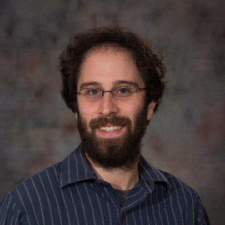 Headshot of a smiling person with dark hair, a beard, glasses, and a dark blue striped shirt