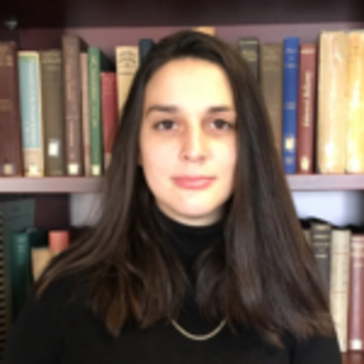 Headshot of a person with long, dark, straight hair wearing a black turtleneck standing in front of a bookshelf