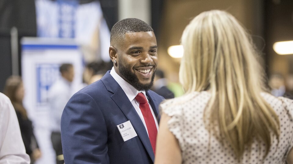 Student and recruiter talking at a career fair with booths and other people behind them