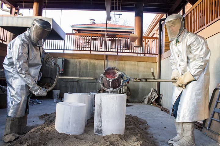 Two people in protective gear pour molten metal into a form.