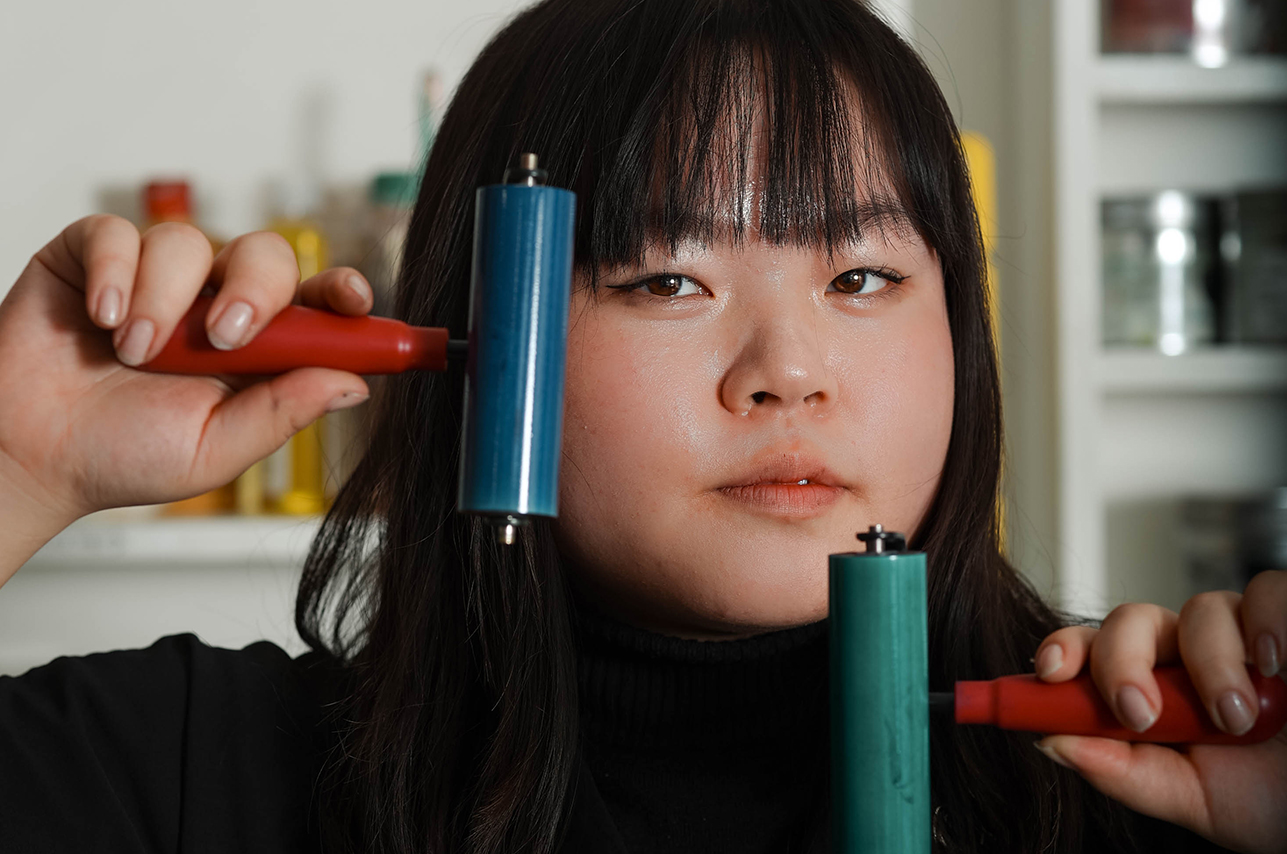 A woman with straight black hair holds to printmaking rollers up in front of her face.