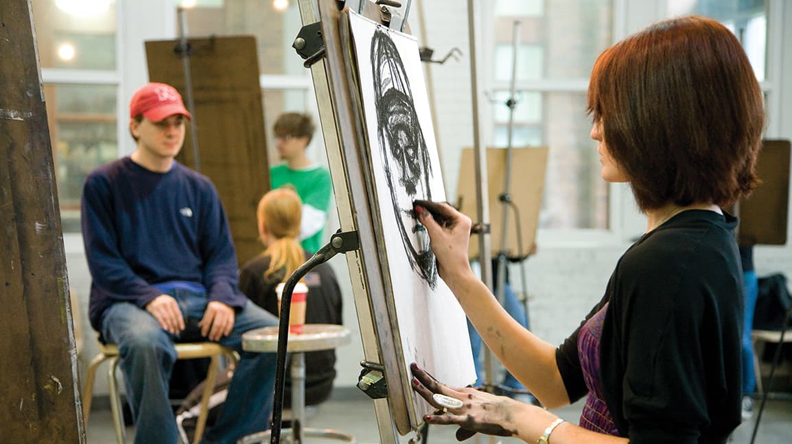 A woman draws while a model sits in the background.