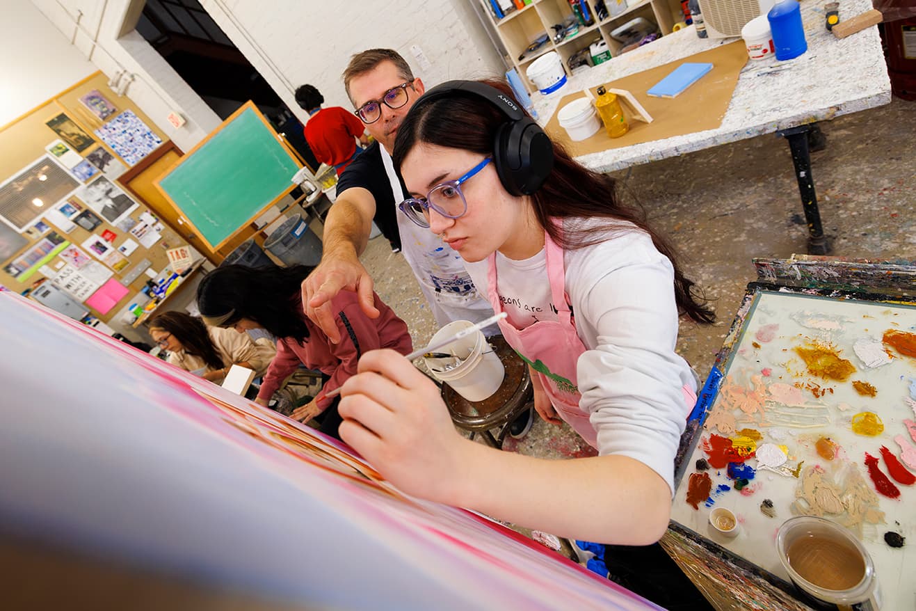 A women wearing headphones holds a brush up to a canvas with a professor pointing to details.