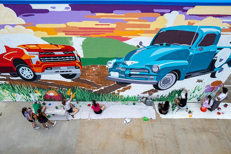 Students working on a giant colorful wall mural of a car and truck.