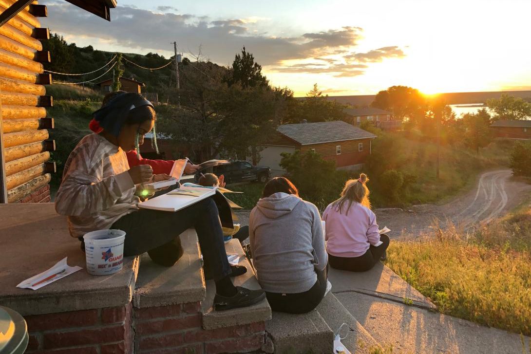 Students sit outside during sun down while working on art projects.