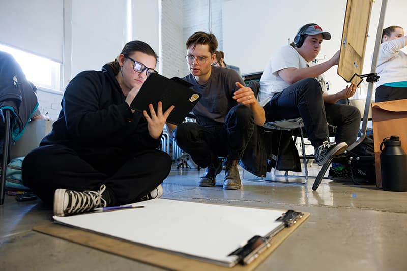 Two people look at reference on a device while sitting before a large pad of drawing paper.