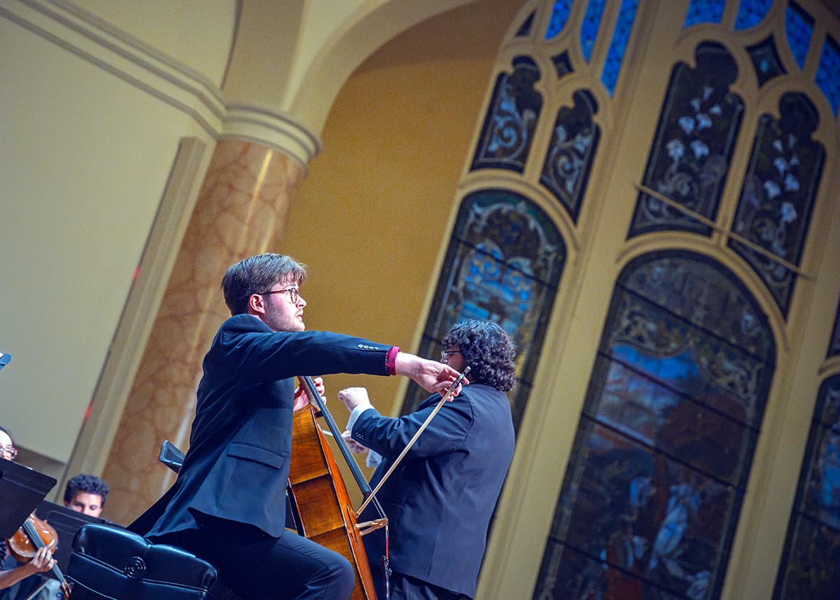 A celloist plays on stage with huge stained glass windows in the background.