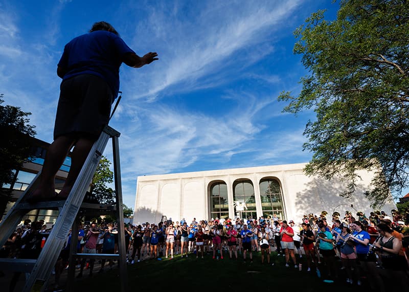 A conductor stands atop a ladder while musicians stand attentively in summer clothes.