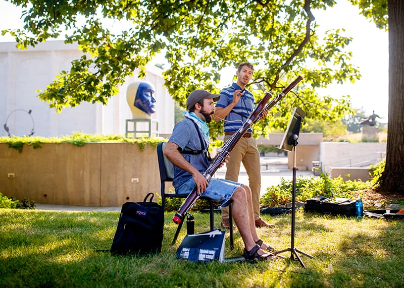 A musician sits on a chair outdoors while a friend helps them practice.