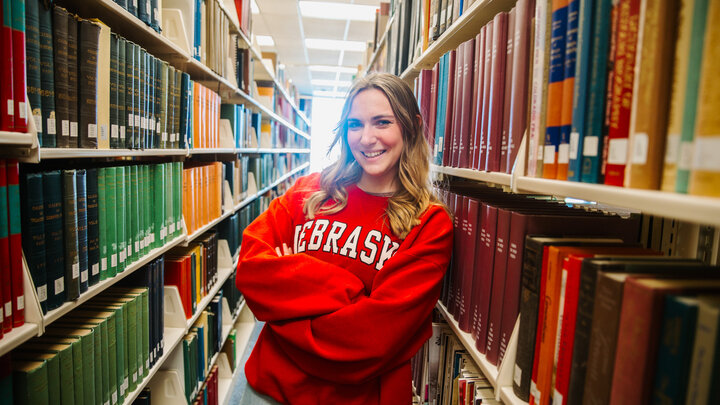 Student wearing red Nebraska sweatshirt leaning against bookshelf in library