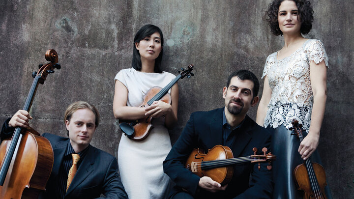 Four musicians with string instruments posing against a textured wall.