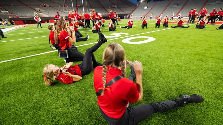 Band members wearing red practice on the Memorial Stadium football field with vibrant green grass at dusk.