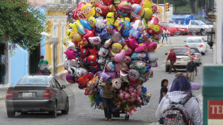 Street vendor carrying colorful balloons on a cart, surrounded by cars and pedestrians.