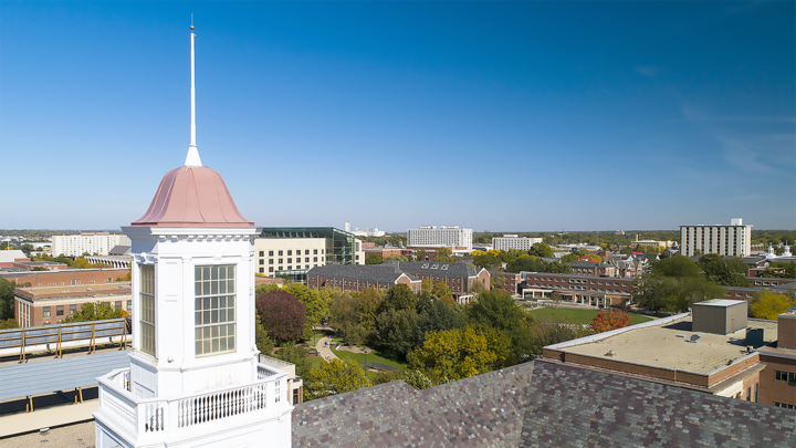Campus view with a white tower, green trees, buildings, and clear blue sky.