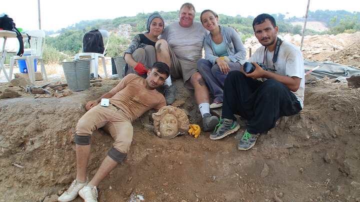 Group of archaeologists sitting near an excavation site with a sculpted head artifact.