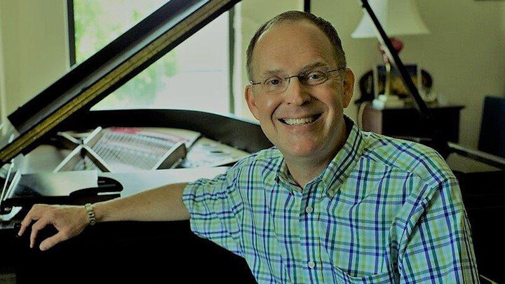 Mark Clinton headshot. He is sitting at a piano in his studio in the former Westbrook Music Building.