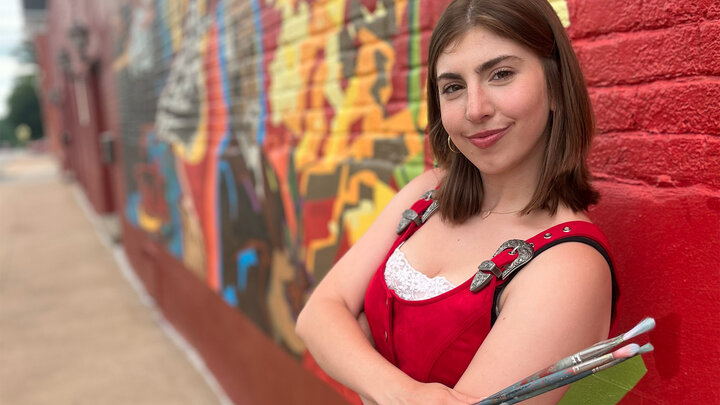 Katie Dia in red top holding paintbrushes, standing in front of a colorful mural on a brick wall.