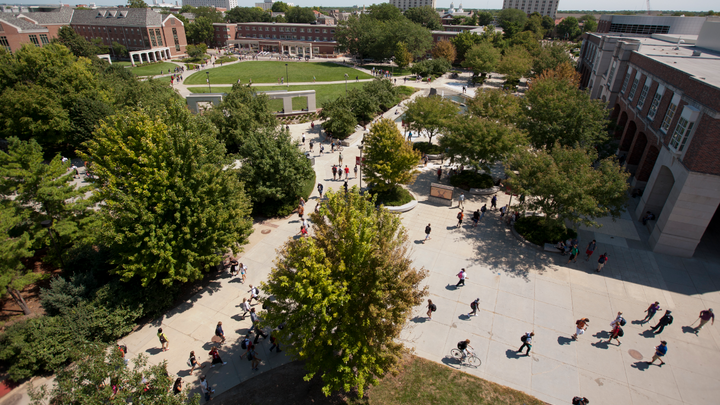 Campus courtyard with students walking, surrounded by trees, buildings, and open grassy spaces.