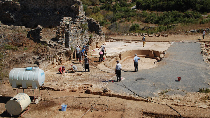 Archaeologists working on ruins with tools, a large container, and scattered vegetation nearby.