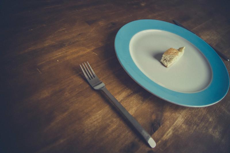 A plate with a blue rim with a small piece of food on it and a fork sit on a wood table