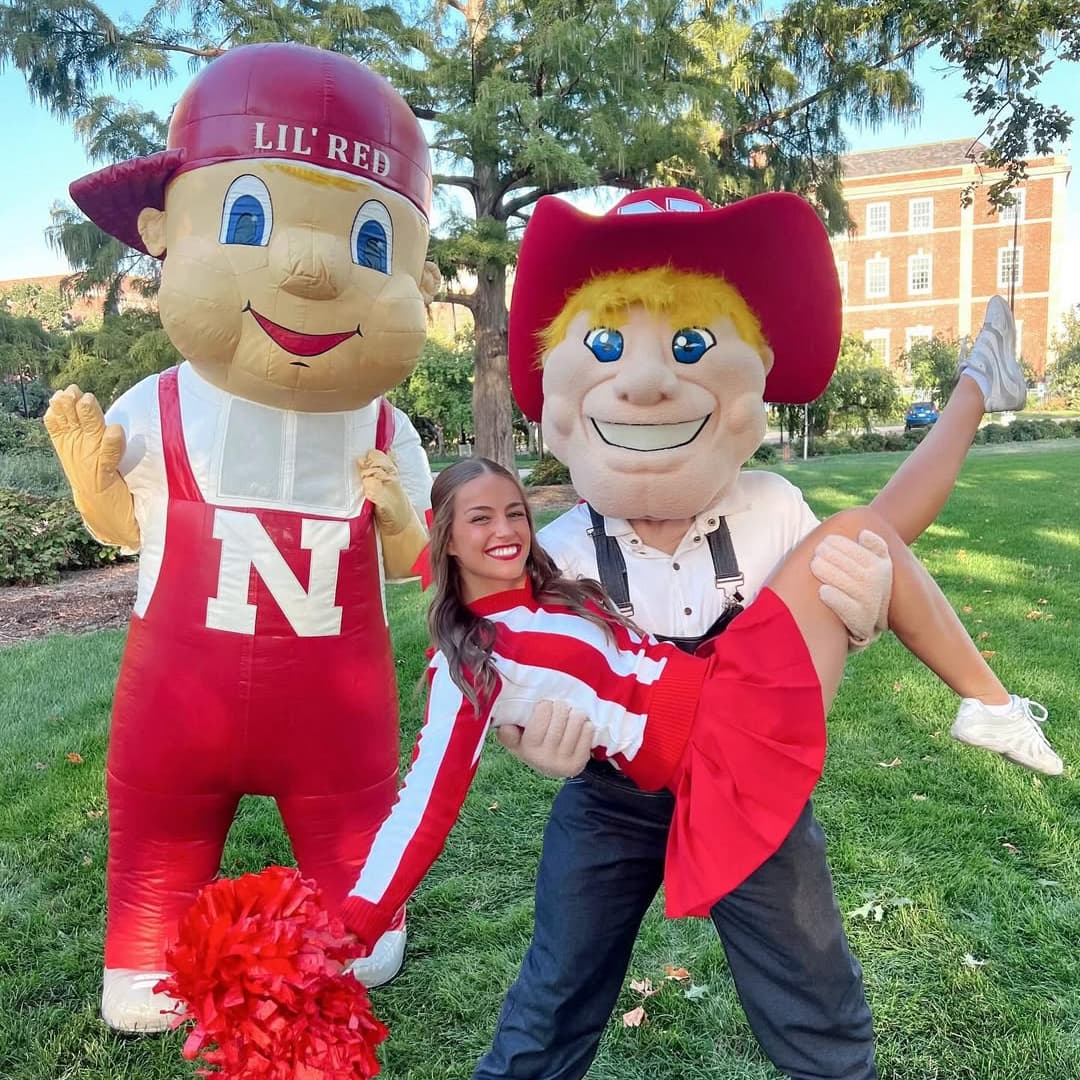 Husker in a cheerleader uniform is held by Herbie Husker who is standing next to Lil' Red.