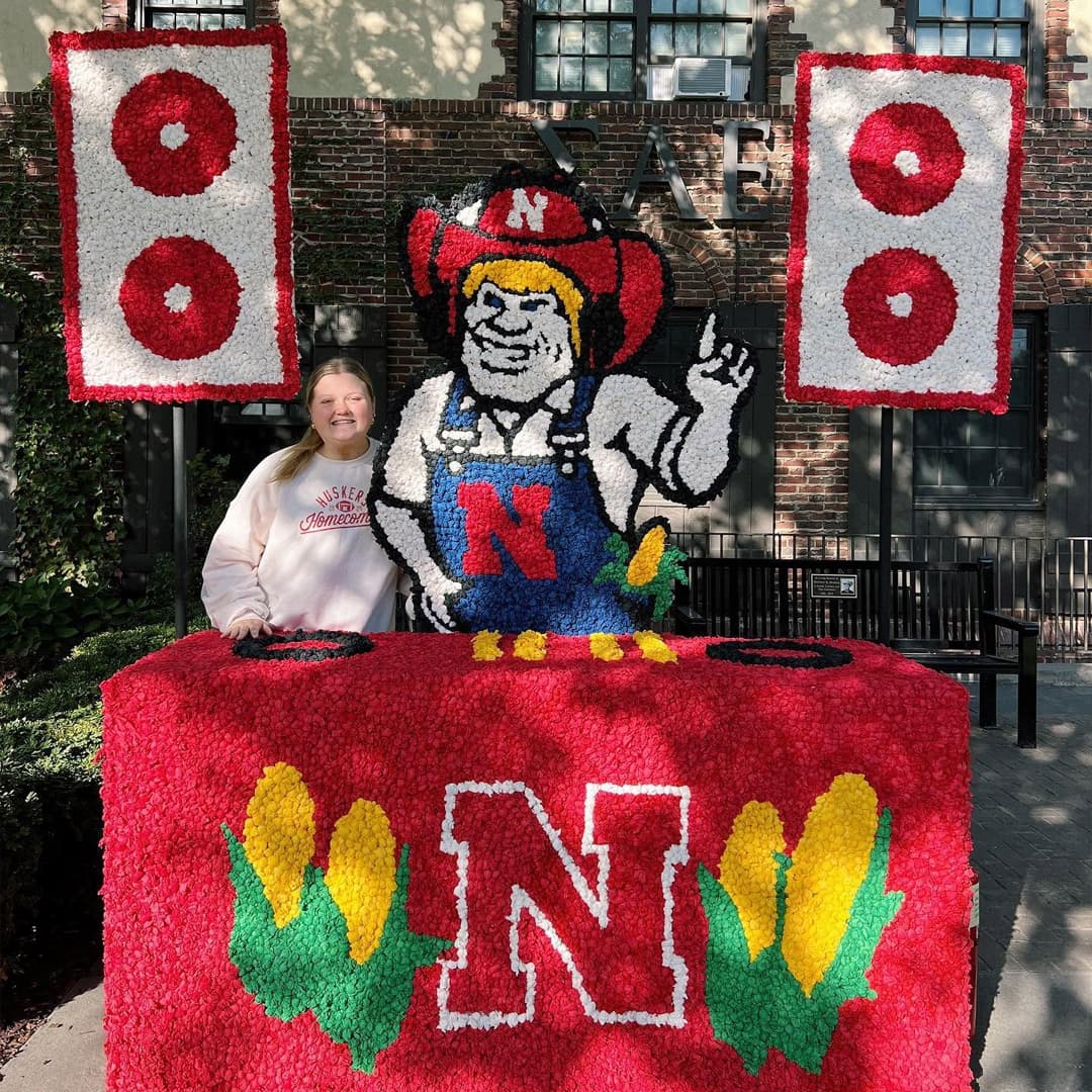 A Husker poses at a Homecoming decoration of Herbie Husker at a DJ table with speakers in the background.