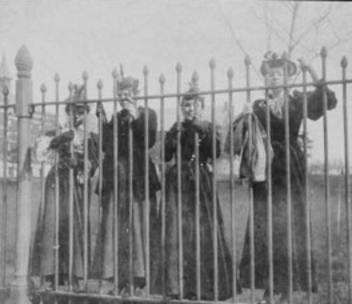 Four women stand behind tall wrought iron fence.